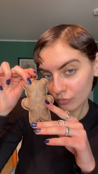 Llavero en forma de oso de cuero color canela con superficie texturizada y detalles de costuras, sostenido por una persona con esmalte de uñas azul y anillos plateados.