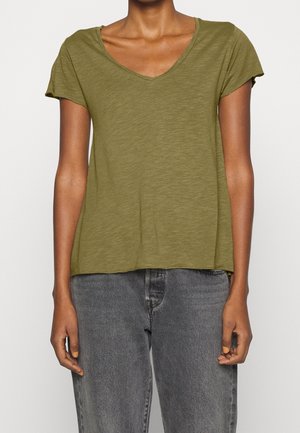 Person wearing an olive green short-sleeve V-neck T-shirt and gray high-waisted jeans, standing with arms relaxed at sides.