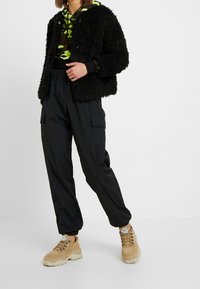 Veste noire en tissu duveteux, sweat à capuche camouflage vert, pantalon cargo noir, baskets épaisses couleur sable. Matières texturées et coupe décontractée.