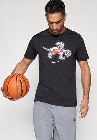 Μαύρο βαμβακερό μπλουζάκι με γραφικό δεινοσαύρου σε κόκκινο χρώμα που κρατάει ένα μπάσκετ και λογότυπο της Nike. Το άτομο φορά γκρι αθλητικό σορτς.