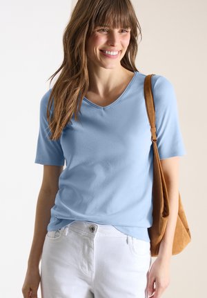 Uśmiechnięta kobieta, ubrana w jasnoniebieską koszulę z krótkim rękawem i białe spodnie, niosąca brązową torbę na ramię po prawej stronie.