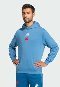 Felpa blu con cappuccio, con emblema di un gallo rosso e strisce bianche Adidas. Realizzata in materiale morbido, con polsini e orlo a costine.