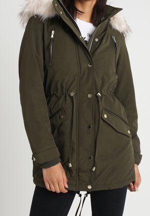 Femme portant une parka vert olive avec une capuche bordée de fourrure, des poches poitrine zippées, de grandes poches à rabat et des cordons ajustables à la taille et à l'ourlet.