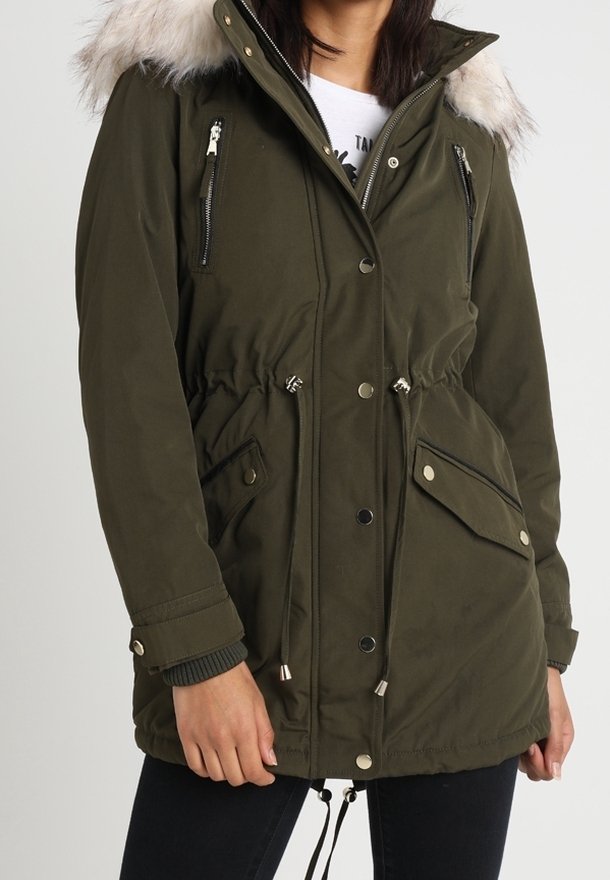 Kobieta ubrana w oliwkowozieloną parkę z kapturem wyściełanym futerkiem, zapinanymi na zamek kieszeniami na piersi, dużymi kieszeniami z patkami oraz regulowanymi ściągaczami w talii i u dołu.