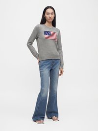 Femme portant un pull gris avec un motif du drapeau américain, un jean évasé bleu et des sandales blanches, debout sur fond blanc.