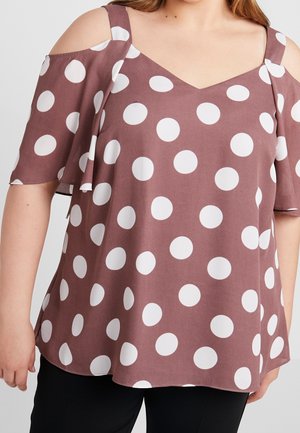 Femme portant un chemisier mauve à épaules dénudées avec de grands pois blancs et des manches volantées, assorti à un pantalon noir.