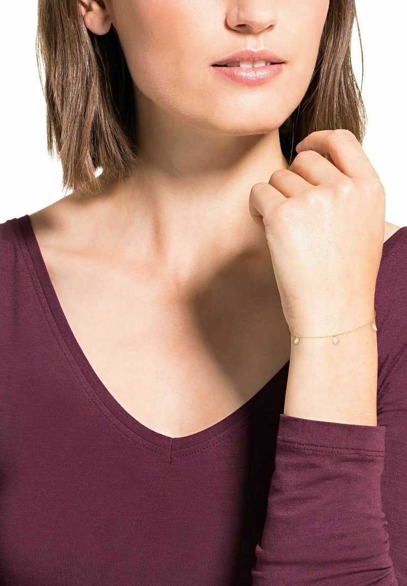 Pulsera de cadena de oro con tres pequeños charms en forma de lágrima. El modelo lleva una blusa de un color burdeos profundo, mostrando la pulsera en su muñeca.