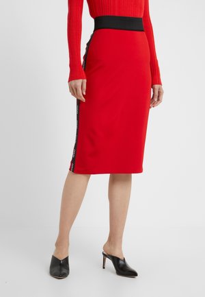 Kobieta w czerwonej spódnicy do kolan z czarnym paskiem w talii oraz czarnymi bocznymi paskami z białym napisem, w połączeniu z czarnymi mules na obcasie.