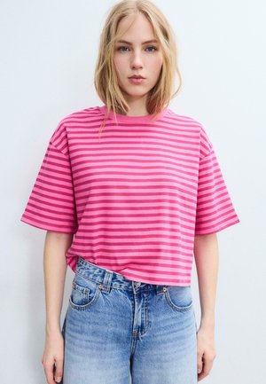 Giovane donna con capelli biondi fino alle spalle che indossa una maglietta a righe rosa e magenta e jeans a vita alta contro uno sfondo semplice.