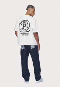 Balta kokvilnas T-krekls ar melnu apļveida grafiku uz muguras. Kombinēts ar tumšām džinsa biksēm, kurās ir zili akcenti un izdrukas.