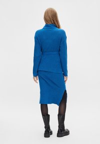 Ensemble en tricot bleu composé d'un pull et d'une jupe avec tissu texturé, manches longues, col montant et ourlet asymétrique. Porté avec des bottes noires massives.