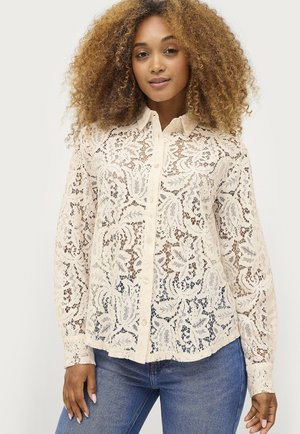 Bluse i lace i creme med blomsterprint, knaplukning foran, lange ærmer og en let buet kant. Parret med mørke jeans.