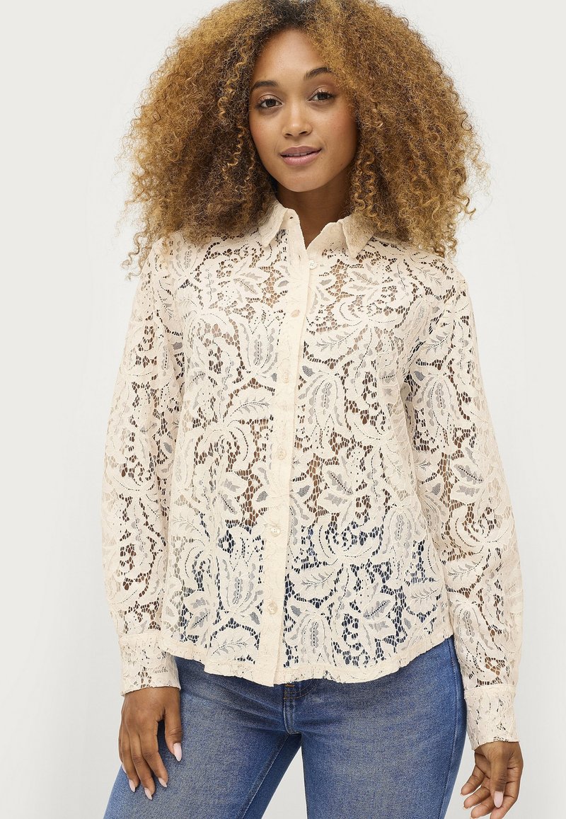 Kanten blouse in crème met bloemenpatroon, knoopsluiting aan de voorkant, lange mouwen en een licht gebogen zoom. Gecombineerd met donkere jeans.