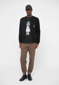 Svart kofta över en svart t-shirt med robotmotiv. Bruna byxor med dragsko; bärs med svarta skor och keps.