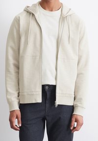 Felpa con zip di colore beige chiaro, dotata di tasca frontale, cappuccio con stringa e polsini a coste. Abbinata a jeans in denim scuro.