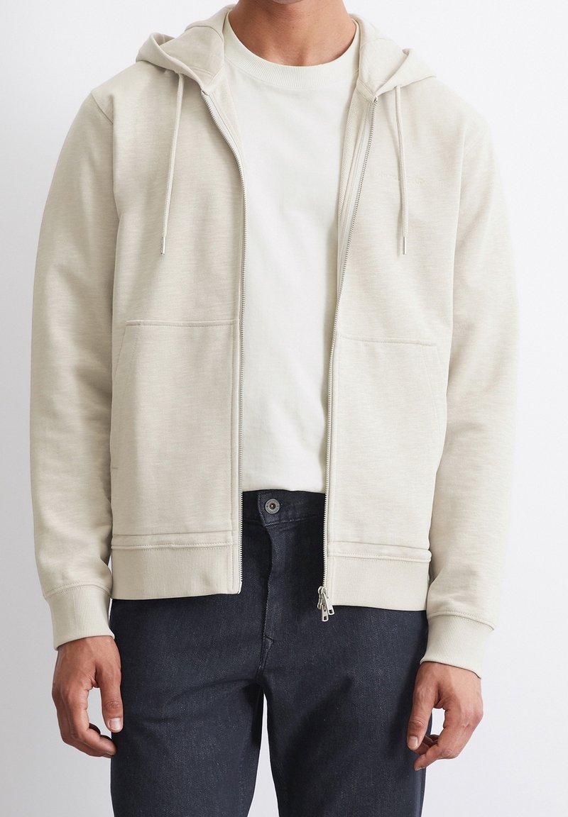 Felpa con zip di colore beige chiaro, dotata di tasca frontale, cappuccio con stringa e polsini a coste. Abbinata a jeans in denim scuro.