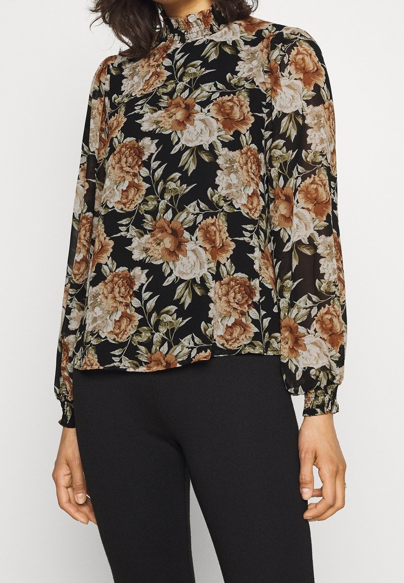 Femme portant un chemisier noir à manches longues avec des motifs floraux marron et blanc et un pantalon noir ajusté.