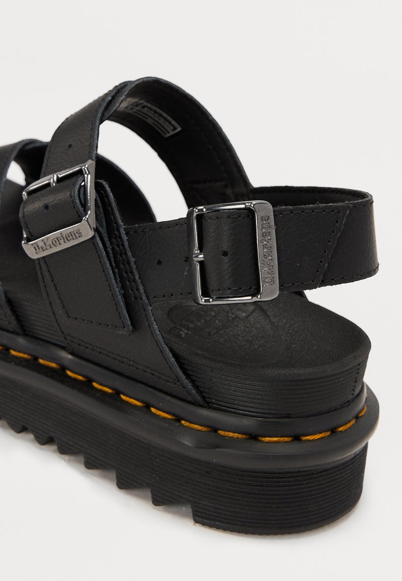 Schwarze Ledersandalen mit verstellbaren Riemen, silbernen Schnallen und einer klobigen schwarzen Sohle mit einem gelben Steppdetail.