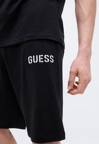 Άνδρας που φοράει μαύρο σορτς με λευκό λογότυπο «GUESS» στο αριστερό μηρό, συνδυασμένο με μαύρο πουκάμισο, στέκεται μπροστά σε ένα απλό φωτεινό φόντο.