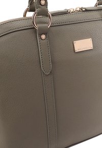 Olijfgroene leren handtas met textuur, goudkleurige metalen ringen, rits en een rechthoekige merkplaat aan de voorkant.