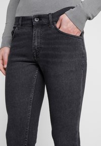 Jeans de mezclilla negra con un diseño ajustado, que cuentan con un bolsillo frontal, cierre clásico de botón y un ligero desgaste en la superficie de la tela.