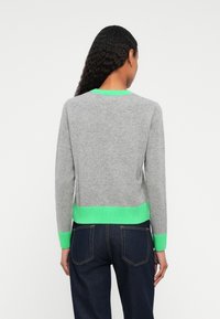 Mulher com cabelo longo e cacheado usa um suéter cinza com detalhes em verde neon e jeans escuros, de costas contra um fundo branco.