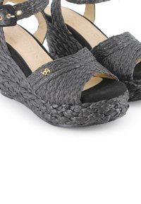 Sandalias de cuña negras tejidas con una textura trenzada, puntera abierta y tira en el tobillo, con un sutil detalle del logo dorado en la parte delantera.