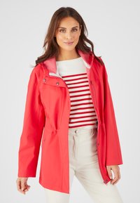 Giacca impermeabile rossa con collo alto, chiusure a zip e a pressione, vita con cordoncino e fodera rosa. Abbinata a una camicia a righe e pantaloni chiari.