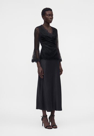 Γυναίκα που φοράει μαύρο δαντελένιο μακρυμάνικο τοπ με μανσέτες καμπάνα και μαύρη σατινέ μίντι φούστα, συνδυασμένο με μαύρα σανδάλια με ψηλό τακούνι.