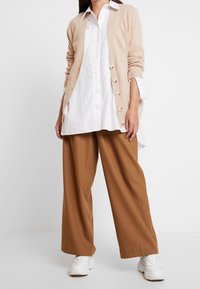 Cardigan de punto beige con grandes botones sobre una camisa blanca de botones, combinado con pantalones de pierna ancha marrones y zapatillas blancas.