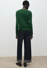 Maglione verde, jeans scuri a gamba larga con polsini a contrasto e scarpe, visti da dietro su uno sfondo neutro.