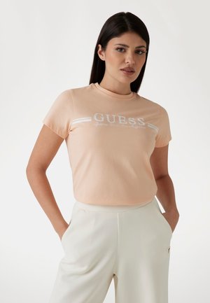 Tyrkysové tričko s krátkym rukávom v broskyňovej farbe s bielou logom "GUESS" a dvoma horizontálnymi bielymi pruhmi na hrudi, vyrobené z mäkkej bavlnenej látky.