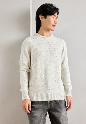 Lyscreme pullover trøje med strukturstrik, der har små mørke prikker, ribbede manchetter og en rund halsudskæring, parret med mørke jeans.