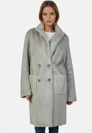 Jeune femme portant un long manteau en fausse fourrure gris clair à double boutonnage avec de grandes poches avant et un large col, debout devant un fond uni.