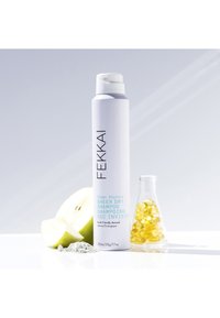 Aérosol blanc avec un texte bleu clair étiqueté "Shampooing Sec Transparent" à côté d'une pomme verte tranchée, d'une bouteille de capsules jaunes et de poudre blanche.