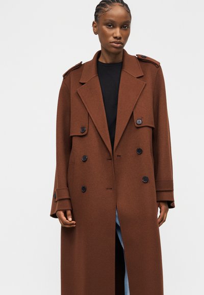 Γυναίκα που φοράει μακρύ καφέ διπλό παλτό με μαύρα κουμπιά, πάνω από μαύρο τοπ και ανοιχτό γαλάζιο τζιν, στέκεται μπροστά σε απλό φόντο.