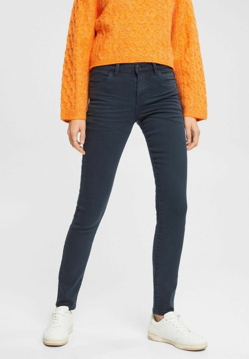 Personne portant un jean skinny bleu foncé, des baskets blanches et un pull en tricot orange texturé, debout les jambes écartées sur un fond blanc.