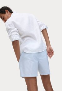 Witte linnen shirt met opgerolde mouwen, gecombineerd met lichtblauwe gestreepte shorts met een ruitpatroon. Model staat met de rug naar de kijker.