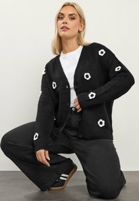 Czarny dzianinowy kardigan z białym haftem w kwiaty, z dekoltem w kształcie litery V, zapinany na guziki z prążkowanymi mankietami i luźnym fasonem. Noszony na białej koszuli.