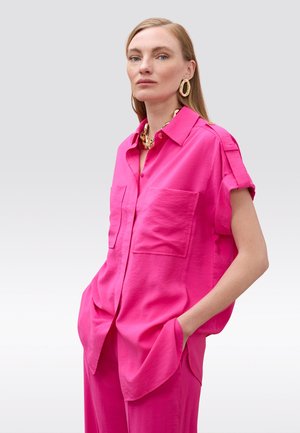 Mujer con camisa de manga corta abotonada de color rosa fuerte y pantalones a juego, accesorios con grandes pendientes de oro y collar, manos en los bolsillos.