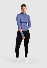 Figurbetonte, nahtlos zu schließende Jacke aus hellblauem Material mit hohem Kragen und Daumenlöchern, kombiniert mit schwarzen Leggings und weißen Sportschuhen.