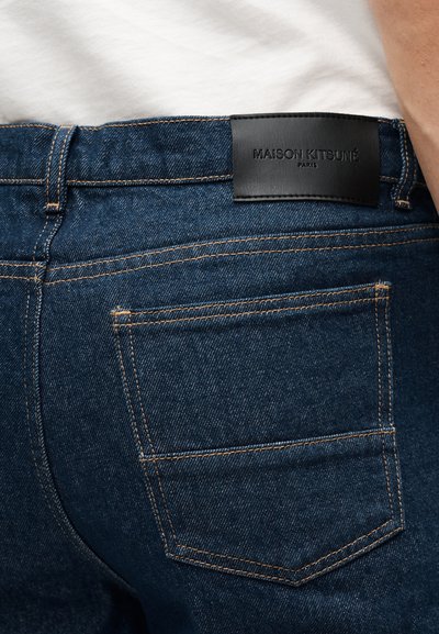 Κοντινή λήψη της πίσω τσέπης μπλε τζιν με πορτοκαλί ραφές και μαύρη δερμάτινη ετικέτα Maison Kitsuné Paris στη μέση.