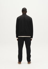 Črna bomberska jakna z rebrastimi manšetami, oblečena čez svetlo srajco, v kombinaciji s temnimi kavbojkami in rjavimi čevlji, pogledano zadaj.