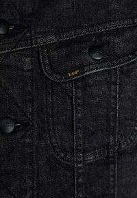 Gros plan sur un tissu denim noir avec une poche cousue, des boutons noirs et une petite étiquette jaune Lee sur le rabat de la poche.