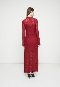 Γυναίκα που στέκεται με την πλάτη γυρισμένη, φορώντας ένα μακρύ κόκκινο δαντελωτό φόρεμα και μαύρα σανδάλια με ψηλά τακούνια, μπροστά σε απλό λευκό φόντο.