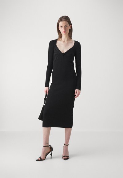 Μαύρο εφαρμοστό φόρεμα με μακριά μανίκια, V-λαιμόκοψη και υφή στην κορυφή. Λείο ελαστικό ύφασμα, μέχρι το γόνατο, συνδυασμένο με ψηλοτάκουνες σandalες με λουράκια.