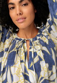 Bluse mit Blumenmuster in Blau, Gelb und Weiß; Ausschnitt mit Schlüssel-Detail und Bindeband; luftiger, strukturierten Stoff mit Falten.