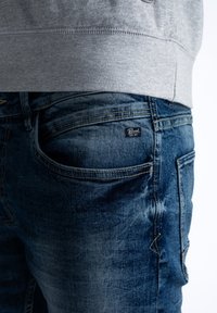 Džíny z denimu v středně modrém odstínu se světlými detaily, s logem na zadní kapse a šedým lemem mikiny viditelným nahoře.