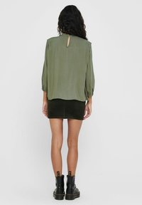 Vrouw met lang zwart haar draagt een olijfgroene blouse, een zwarte minirok en zwarte enkellaarsjes, staande met het gezicht naar een effen lichte achtergrond.