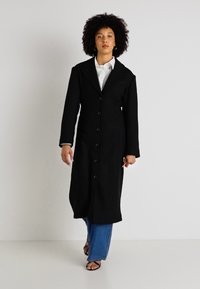 Dlouhý černý kabát s límcem, který má knoflíkovou přední část a boční kapsy. Materiál vypadá texturovaně a teple, doplněný modrými džíny.